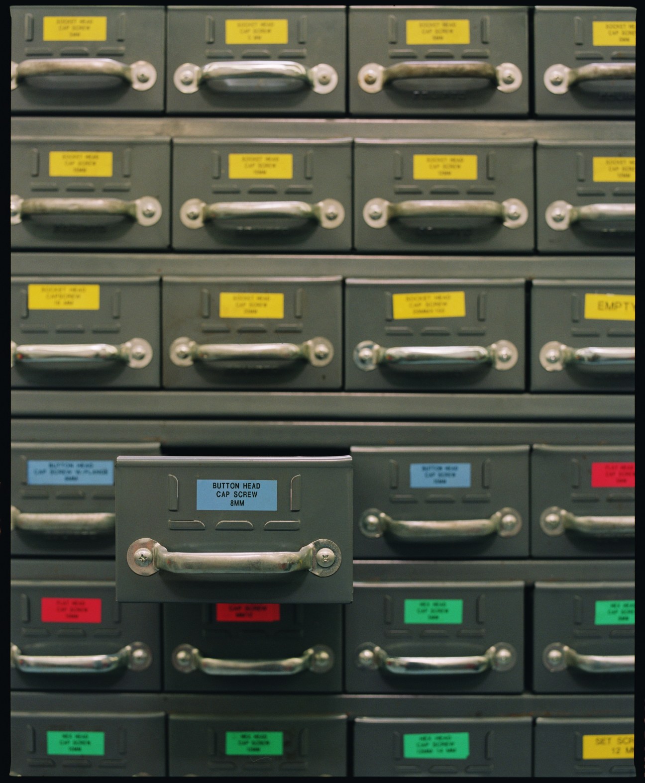 Organized toolshed wall with dark gray storage drawers labeled with yellow, blue, red and green tags, silver handles and an open drawer displaying button head cap screw contents.