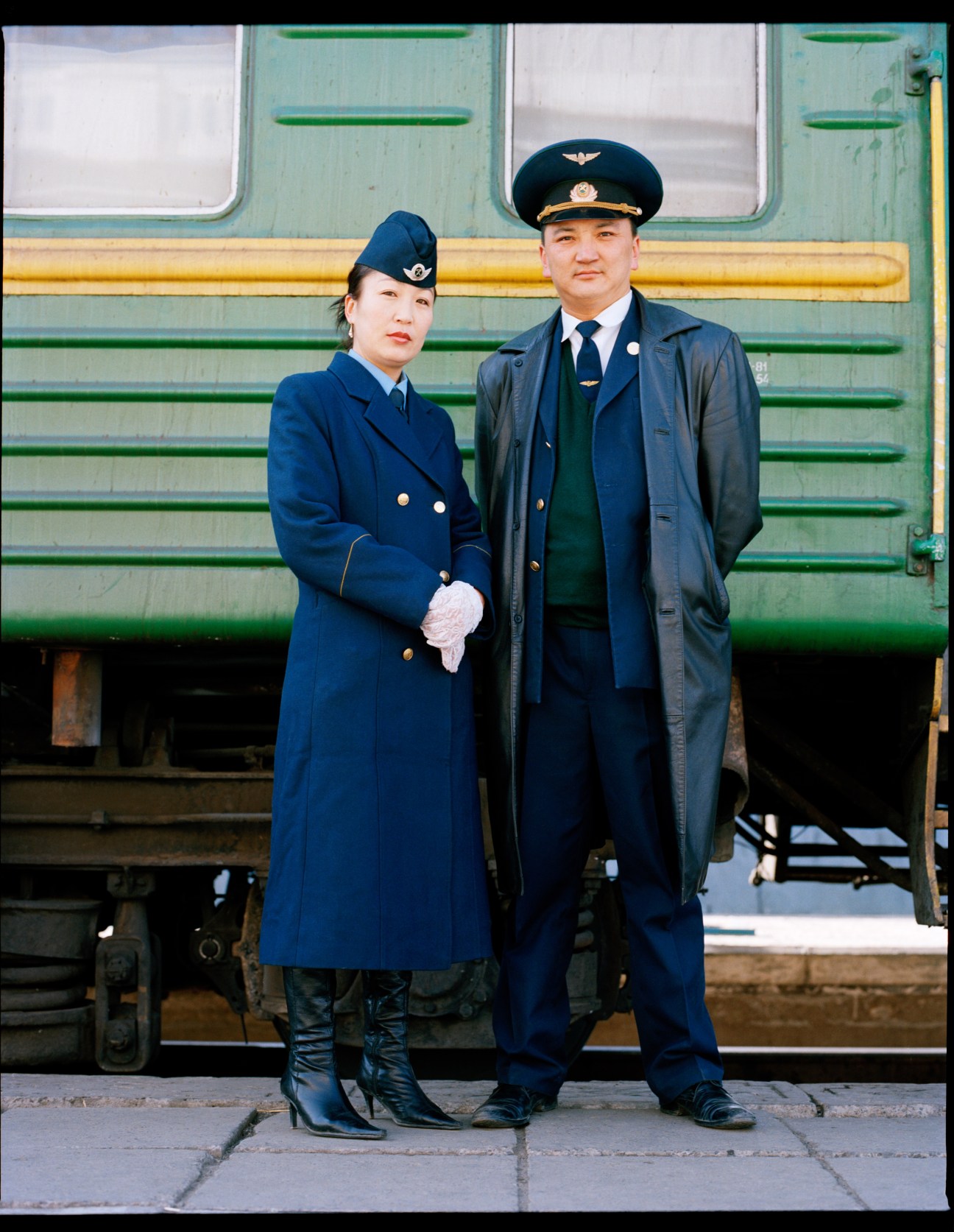 Two uniformed train guards in blue uniforms and caps standing in front of a green and yellow train at Ulan Bator's railway station.