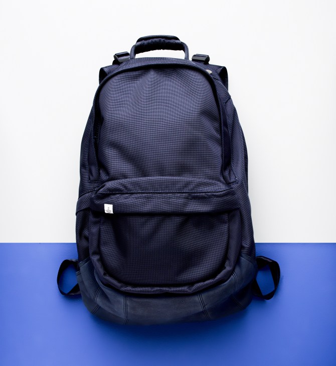 Navy blue Visvim backpack with bullet-proof nylon material and elk leather base, displayed against white and blue background.