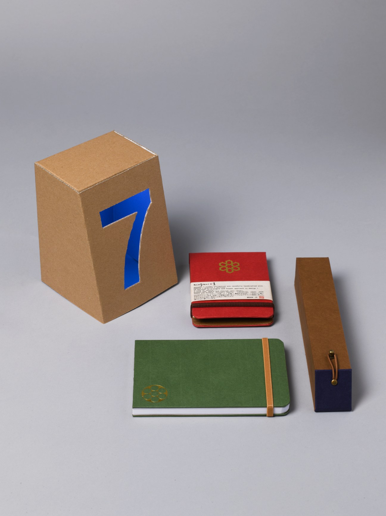 Awagami stationery collection including a cardboard box with blue number 7, red notebook with gold crest, green notepad with elastic band, and brown pencil case.
