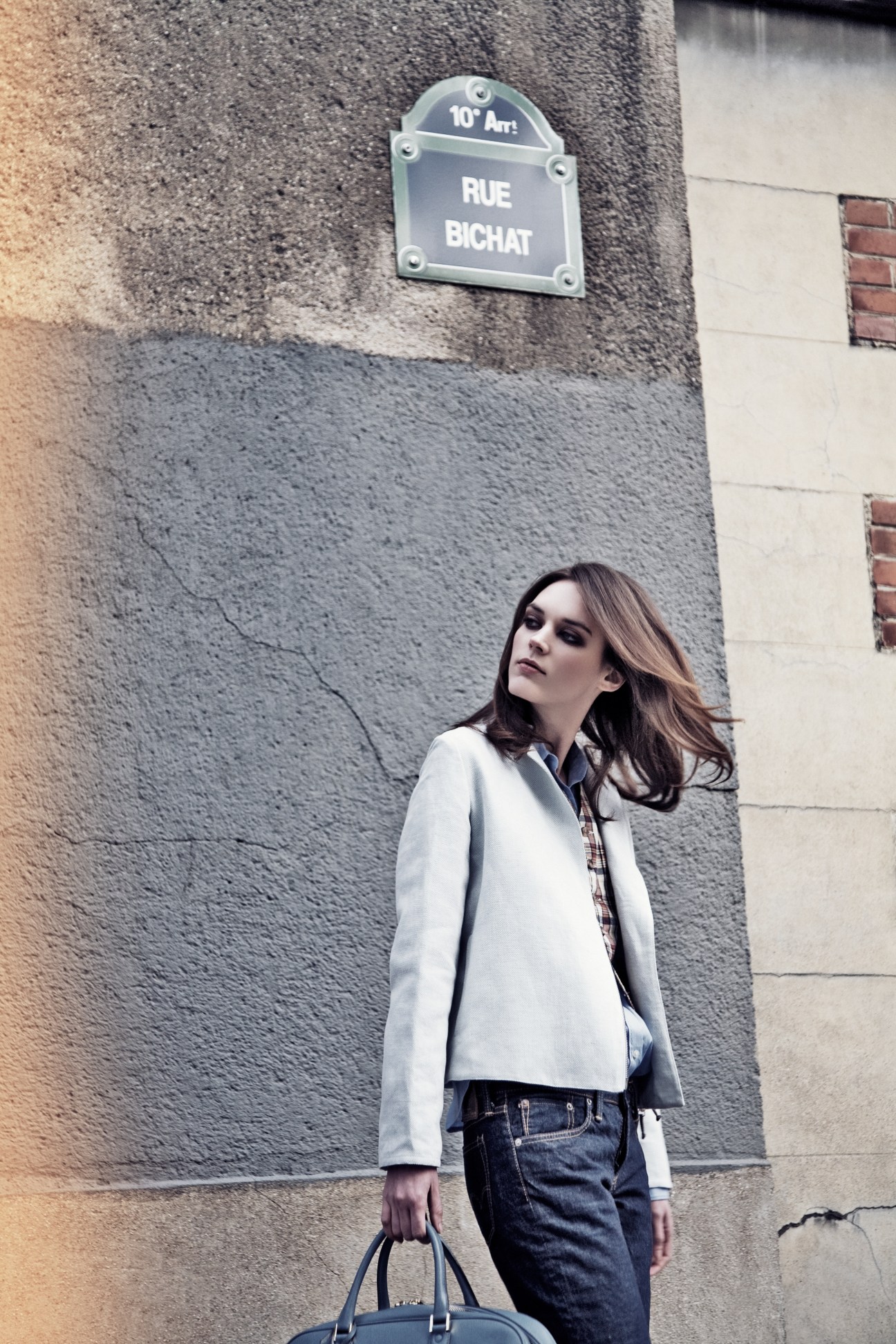 Model wearing a light blue jacket and jeans stands against a Parisian building wall near Rue Bichat street sign.