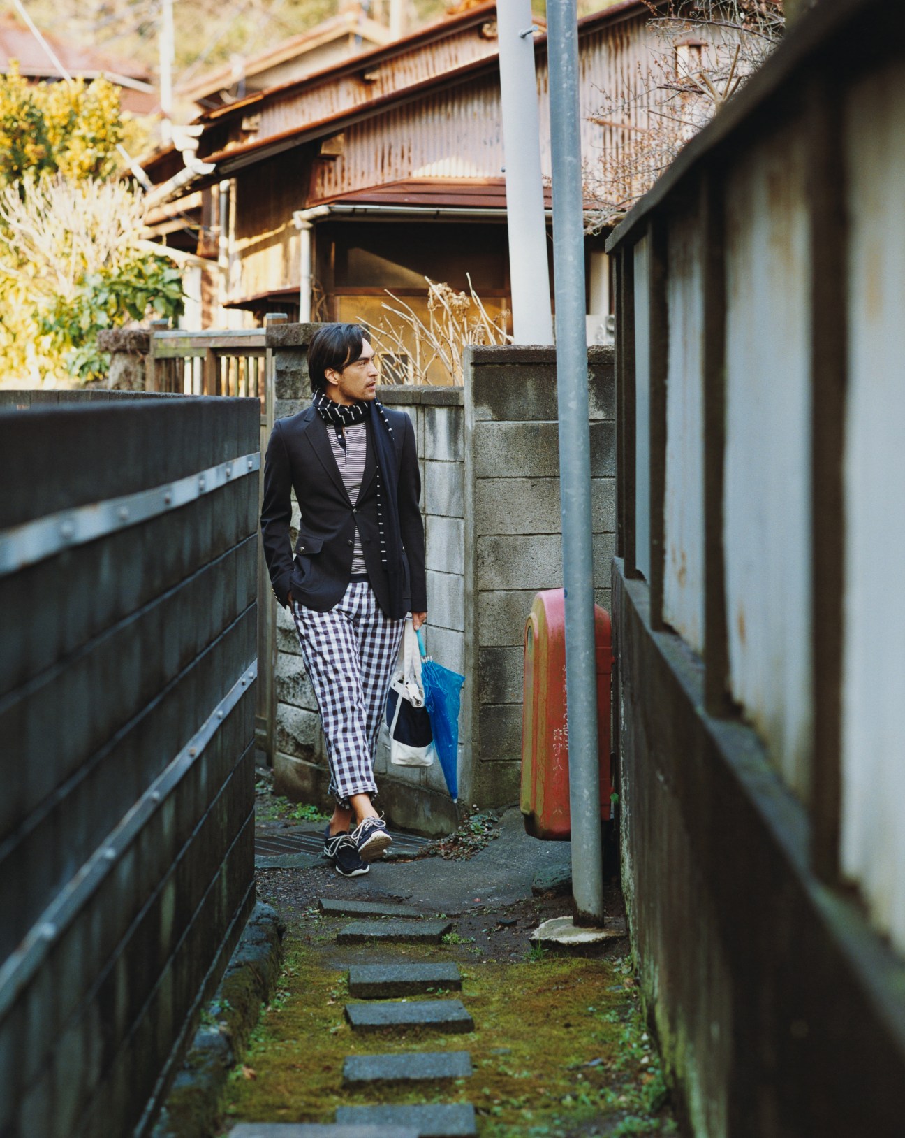 Model wearing a Mackintosh raincoat with Prada layers and Visvim jeans, walking through a narrow alley in Hayama.