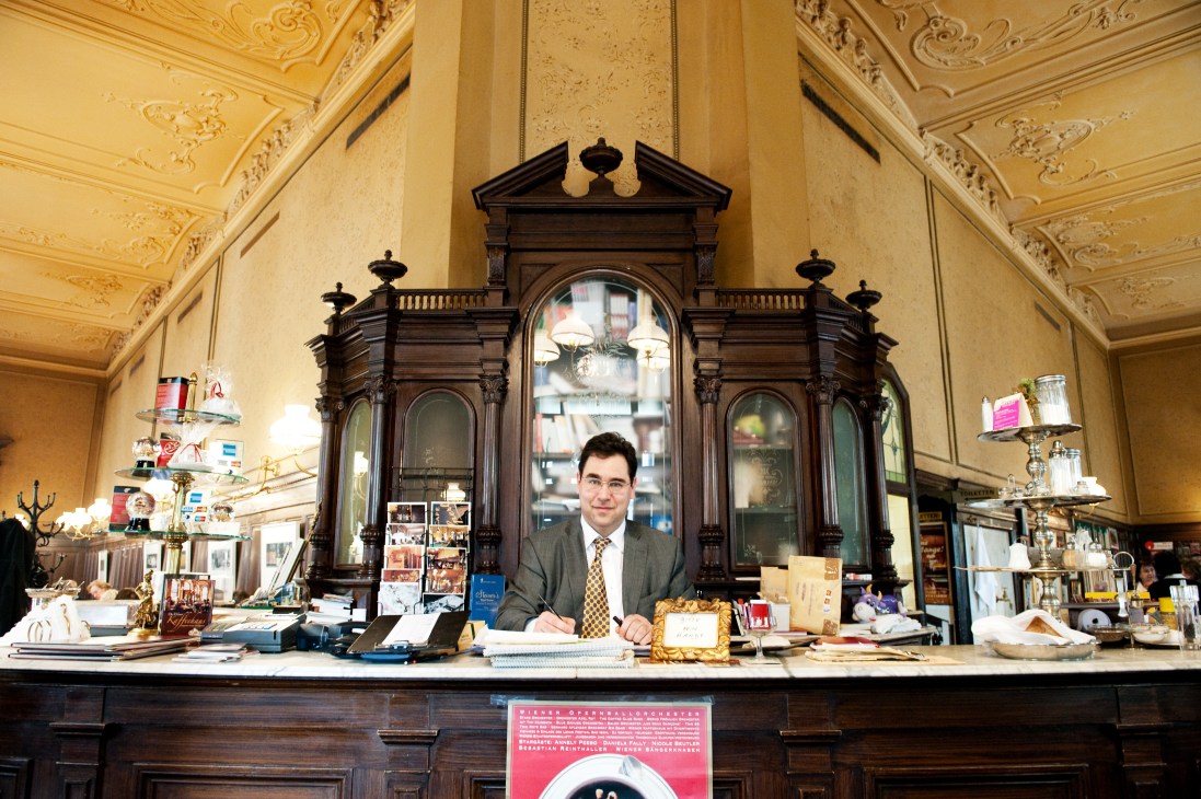 Man in grey suit standing behind counter at Das Möbel furniture shop with ornate ceiling and dark wooden display cabinet.