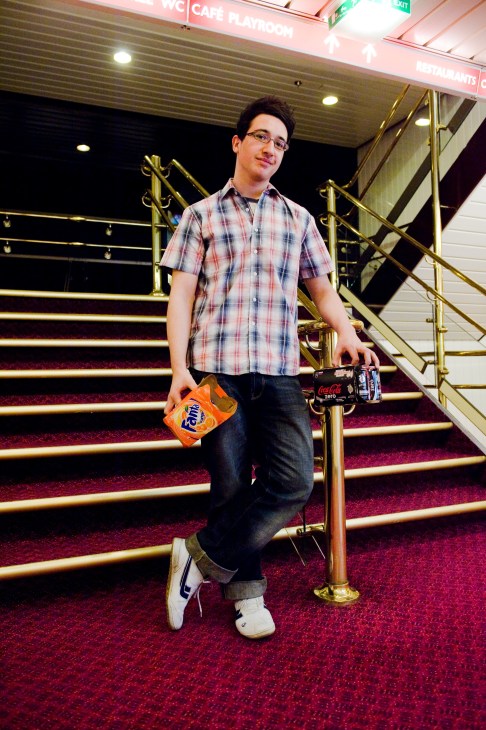 Young man in plaid shirt holding orange Fanta and Coca-Cola Zero on ferry stairs.
