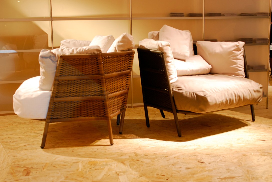 Outdoor furniture display featuring woven rattan chairs and cushioned seating against a golden glass backdrop.
