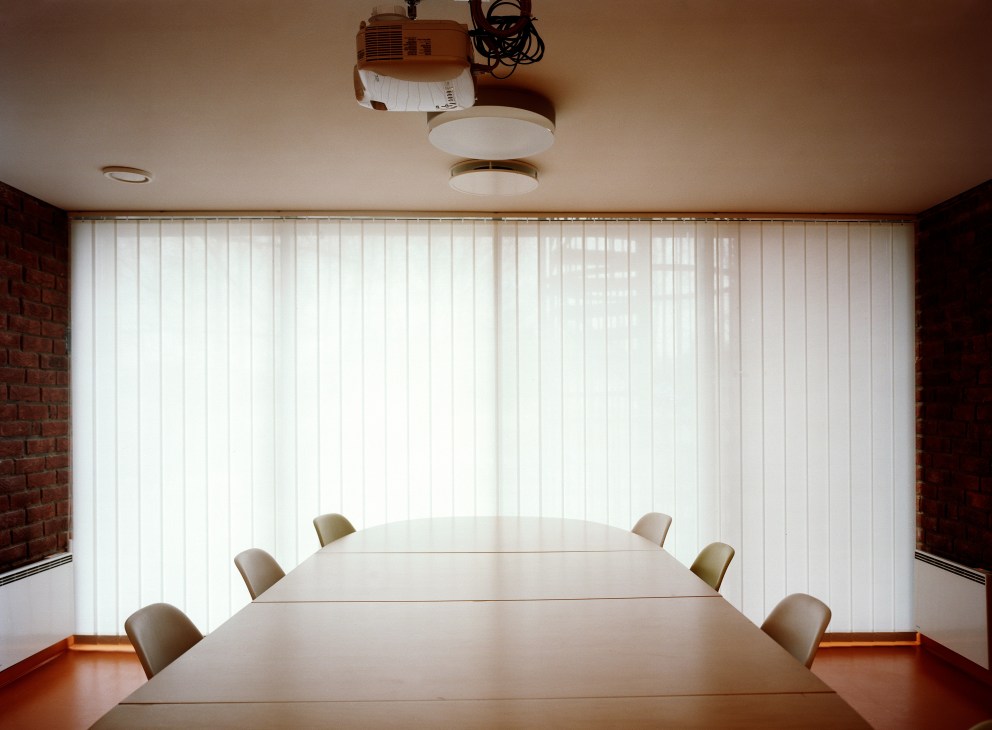 A minimalist meeting room with a long white table, beige chairs, and floor-to-ceiling vertical blinds overlooking natural light.