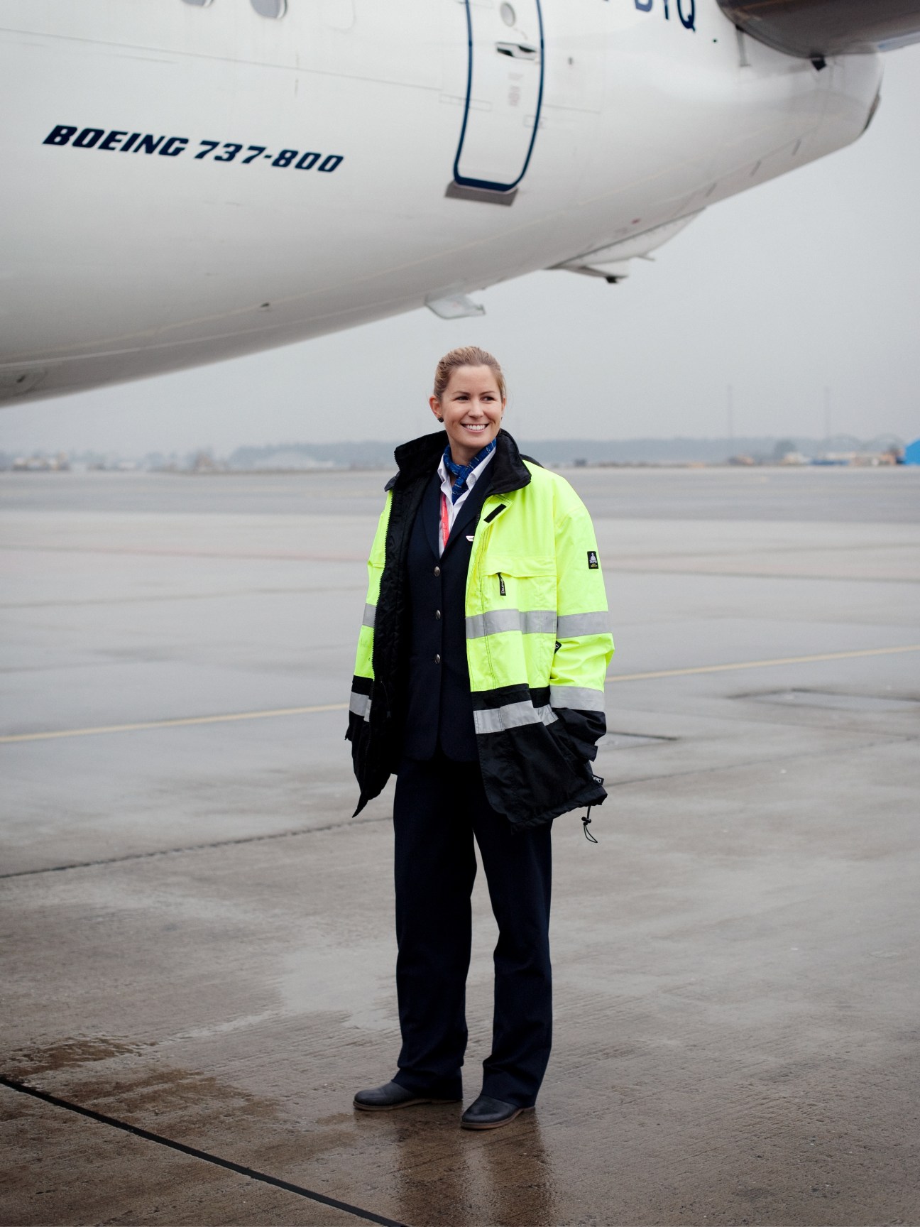 Woman in neon yellow and black safety vest standing on airport tarmac in front of Boeing 737-800 aircraft.