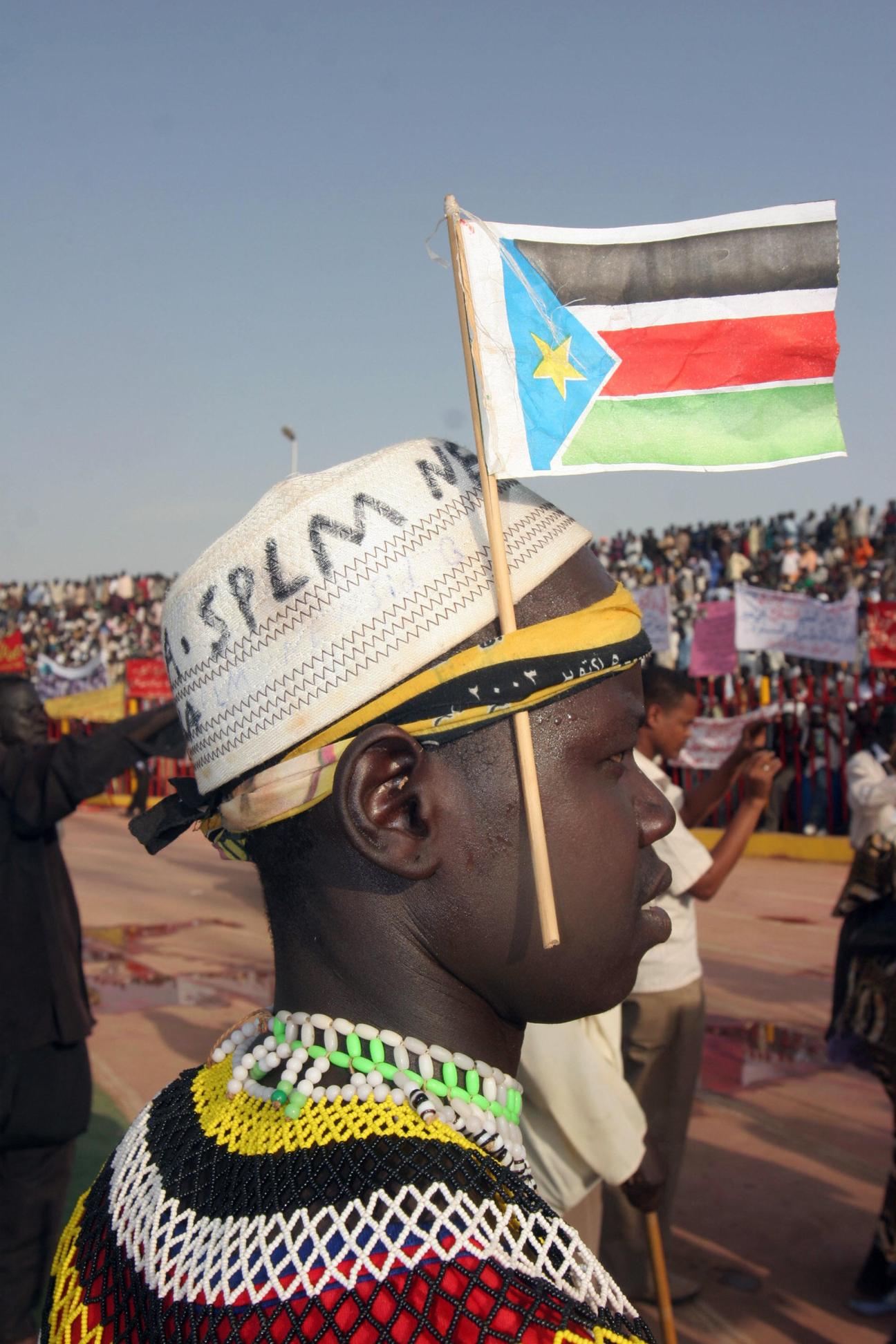 Man in traditional beaded attire holding South Sudanese flag at a rally.