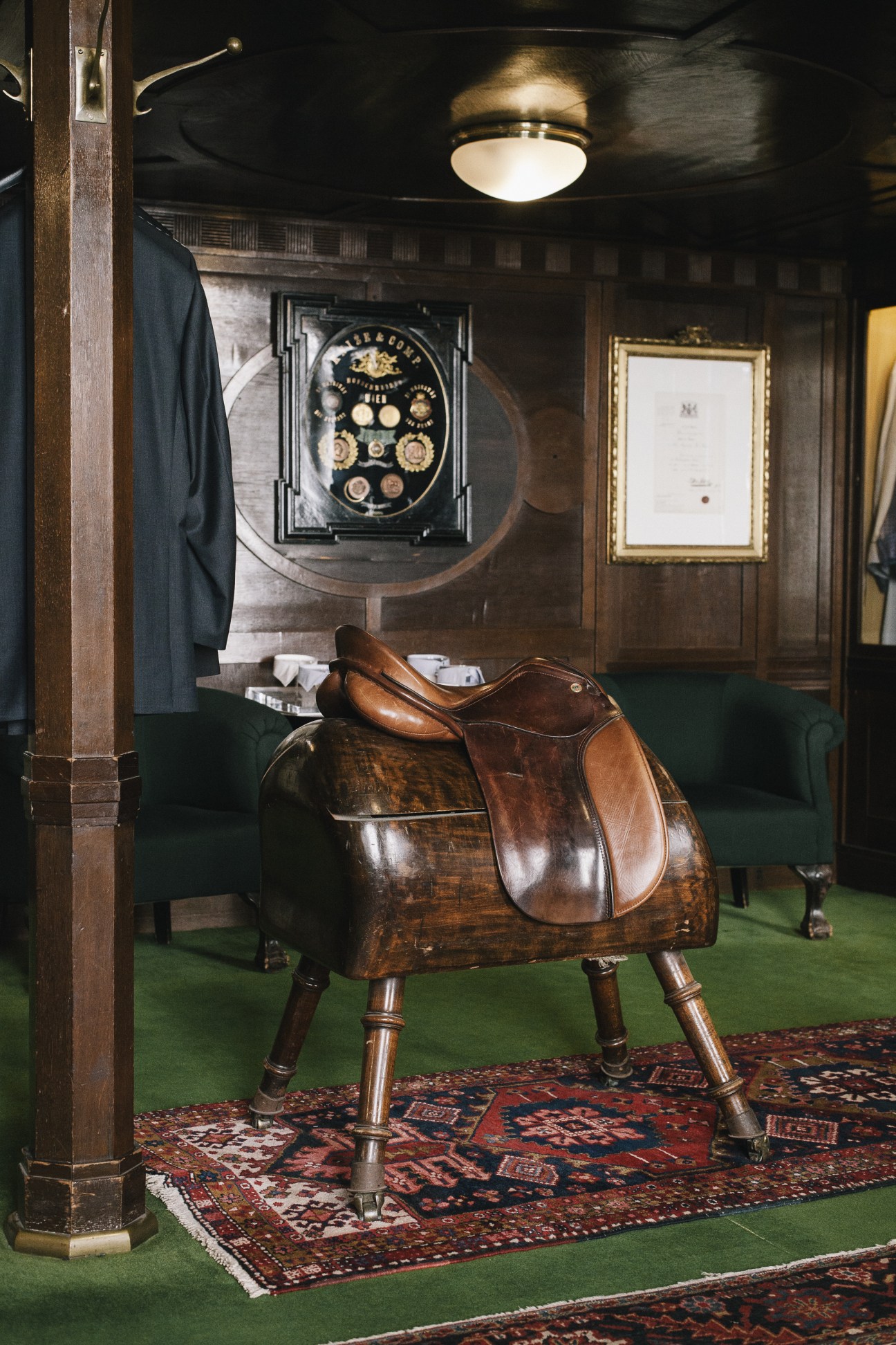 Interior of Knize tailor shop showing a leather saddle chair, framed certificate, astronomical clock, and hanging navy suit jacket.