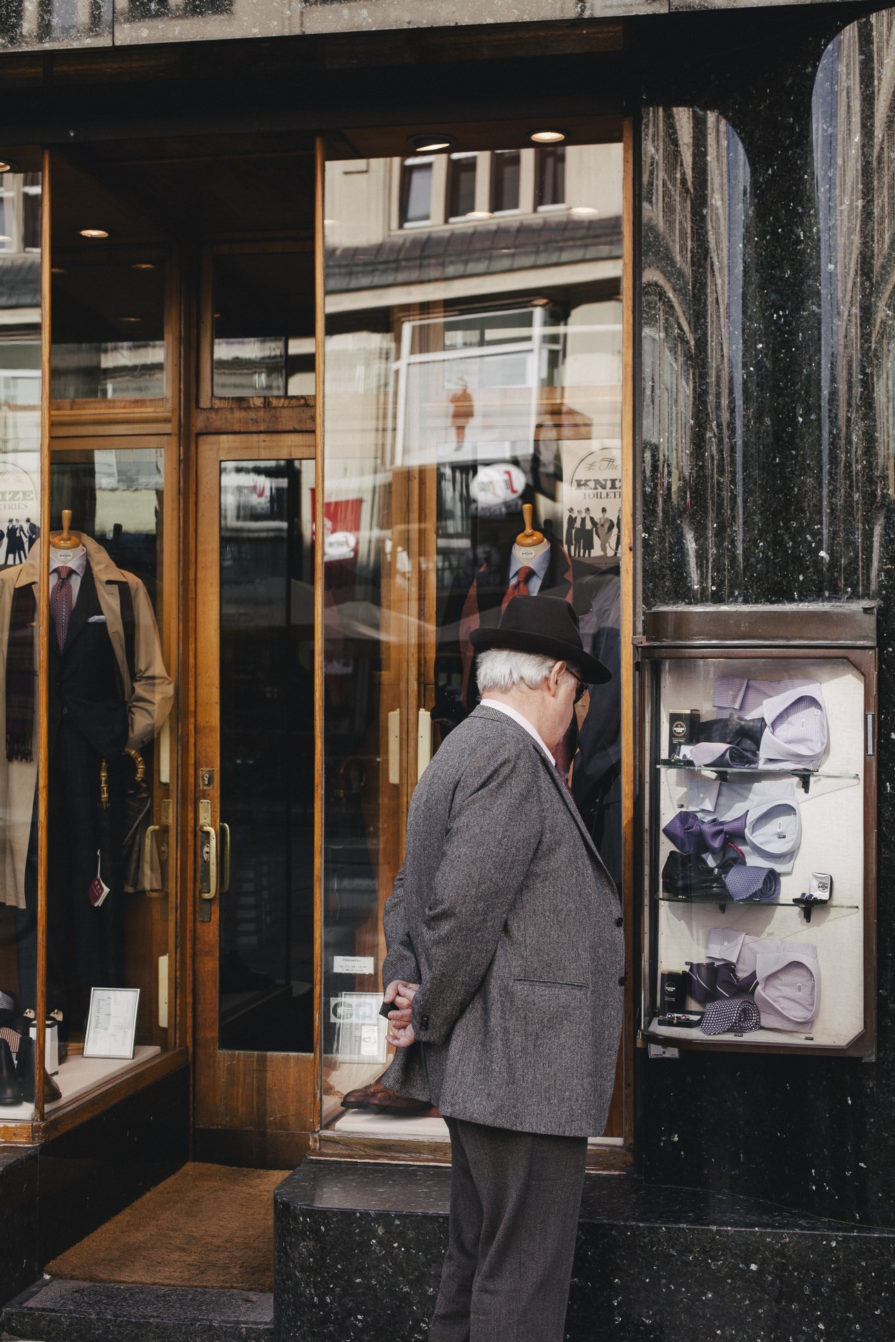 An elderly man in a grey suit and fedora hat stands at the entrance of Knize, a traditional Viennese menswear shop with wooden frames and display windows.