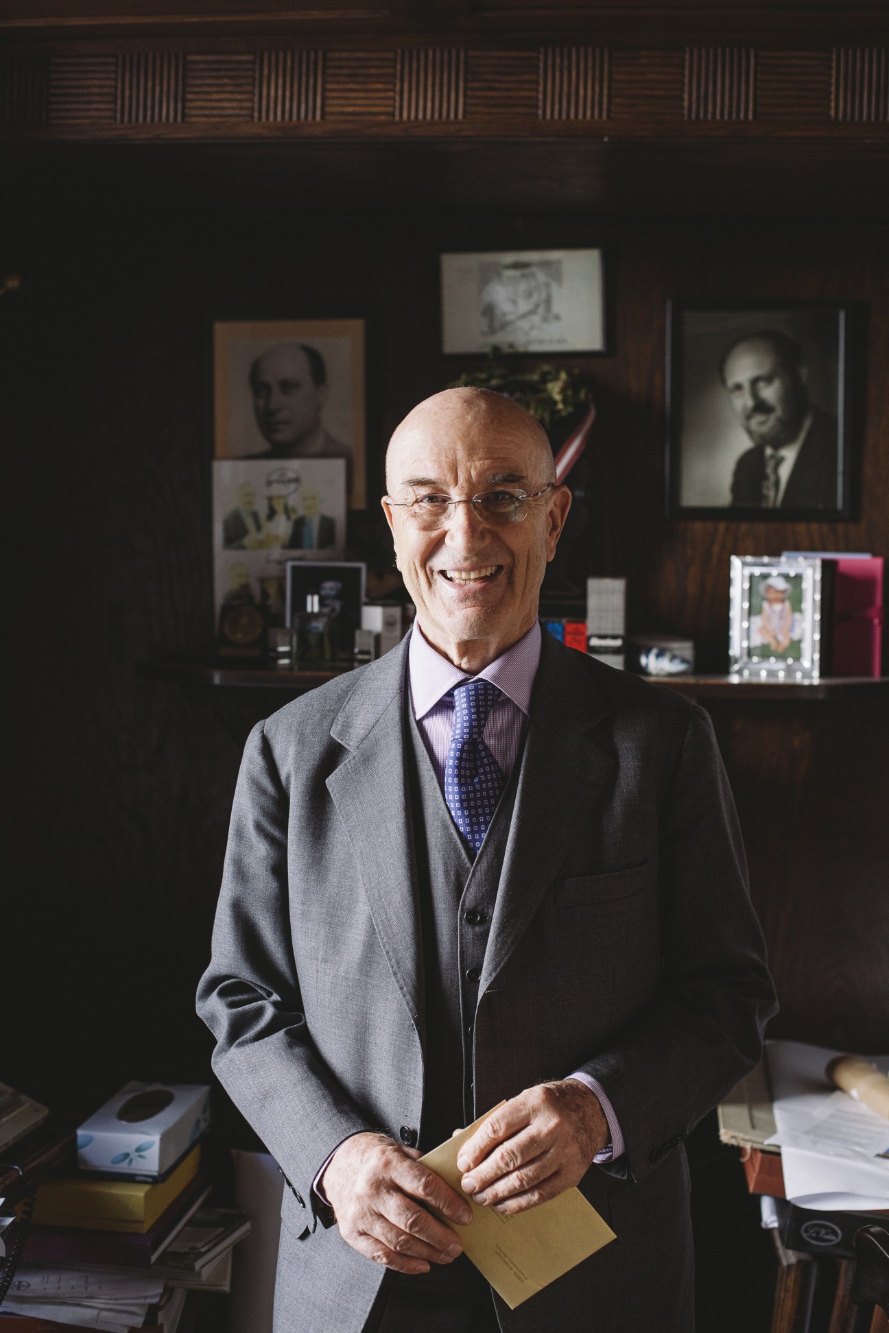 Man in gray suit and blue tie standing in wood-paneled office with historical portraits on wall behind him.