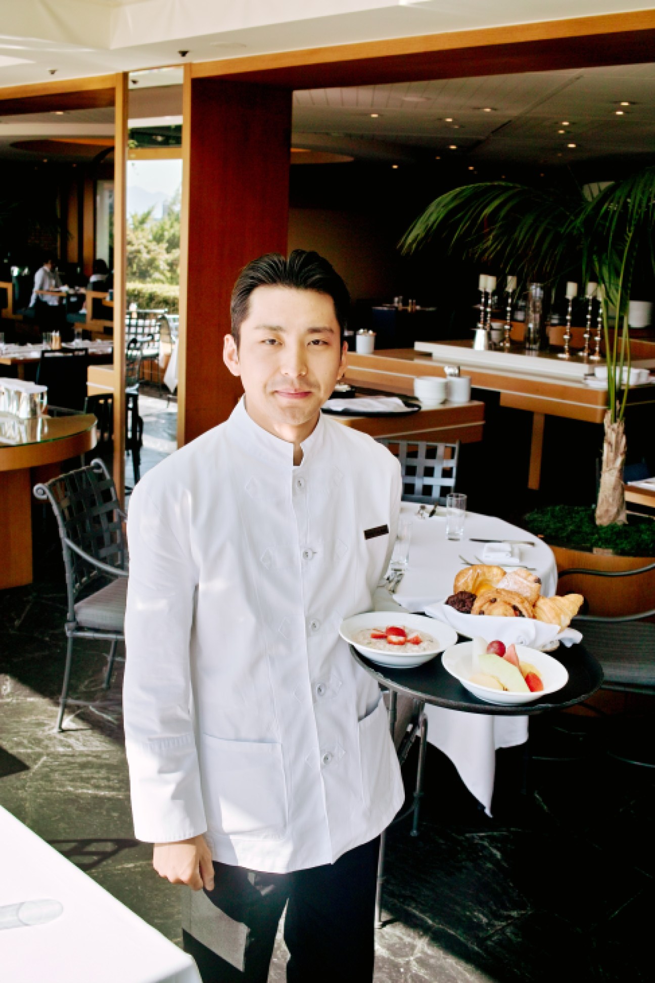 Server in white jacket holding breakfast tray in modern hotel dining room with wood accents and plants.