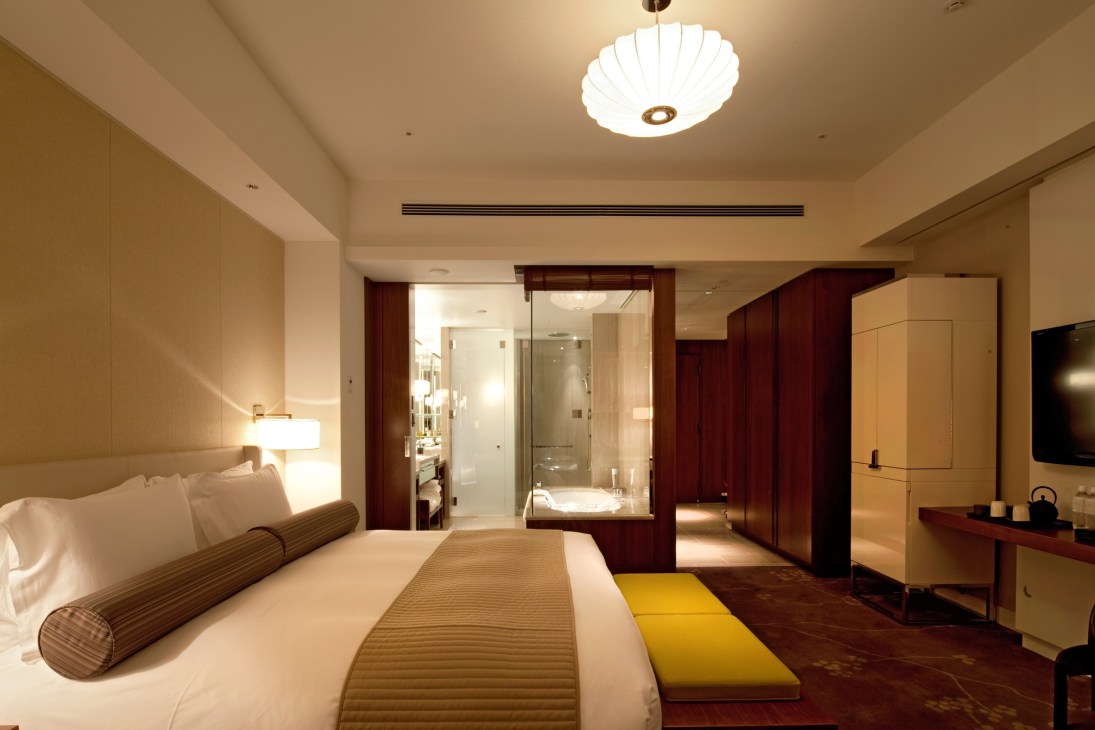 Modern hotel room with bed, wooden accents, glass bathroom enclosure and contemporary lighting.