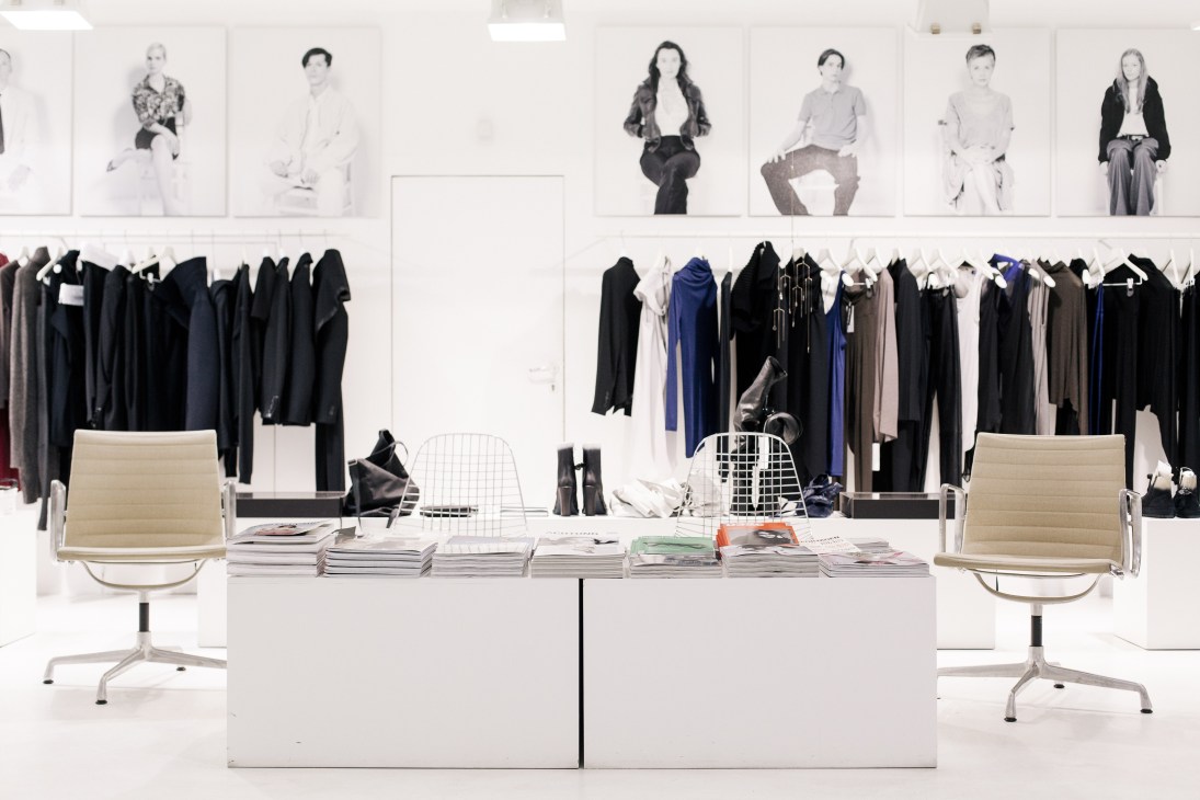 Minimalist fashion boutique interior with black and neutral clothing on racks and portrait photos displayed on white walls.