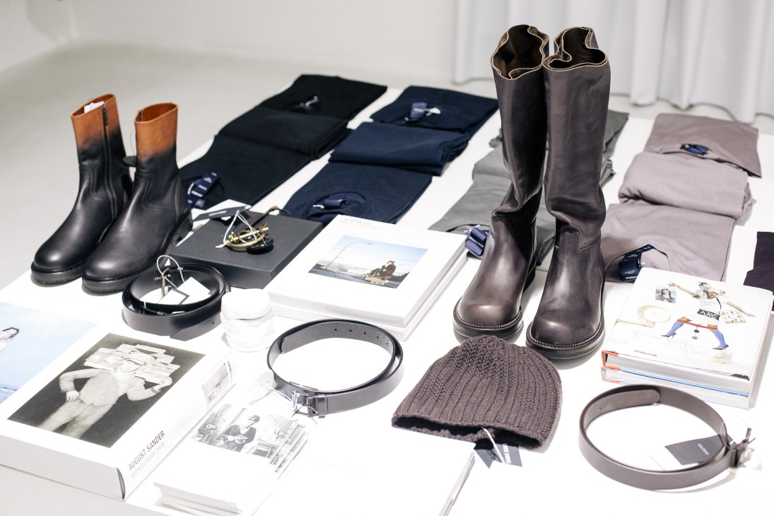 Fashion and accessories display including leather boots, belts, knit beanie, folded clothing, and product catalogs on white surface.
