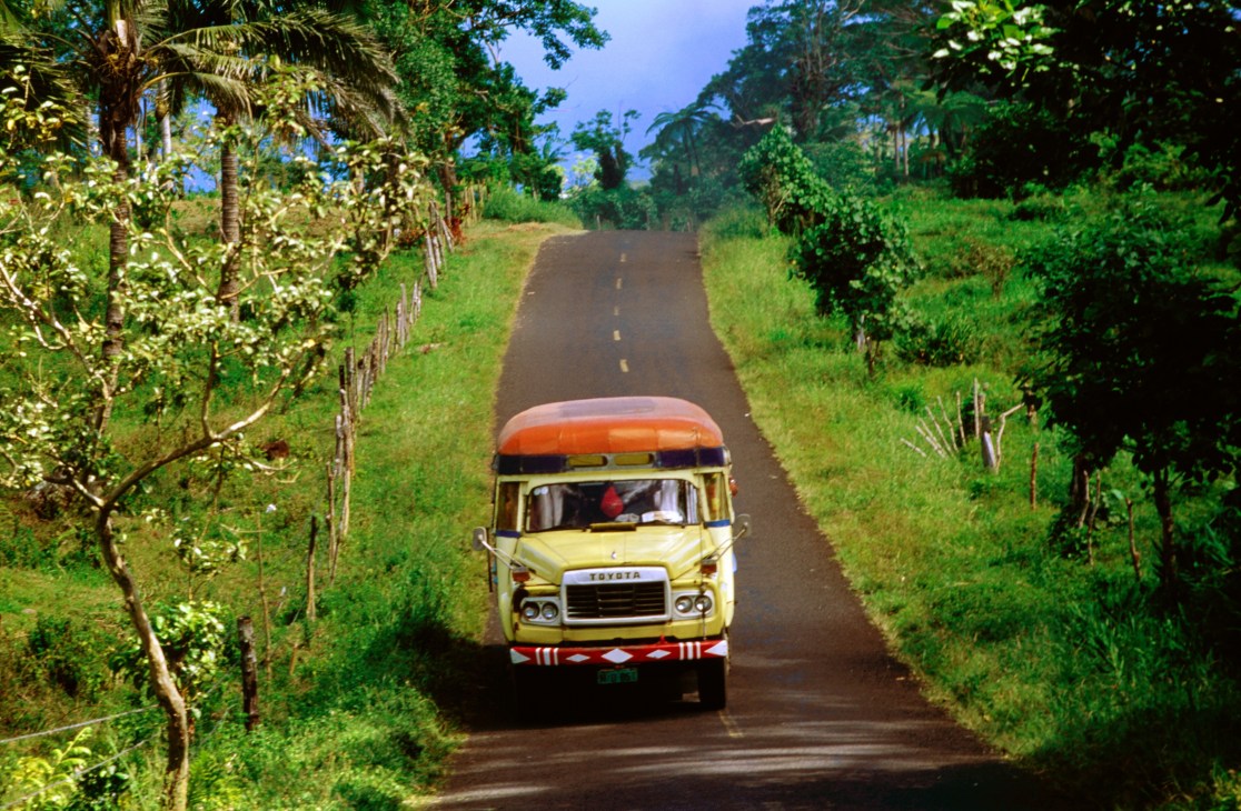 Colorful yellow and orange truck driving on a rural road lined with tropical vegetation in Samoa.