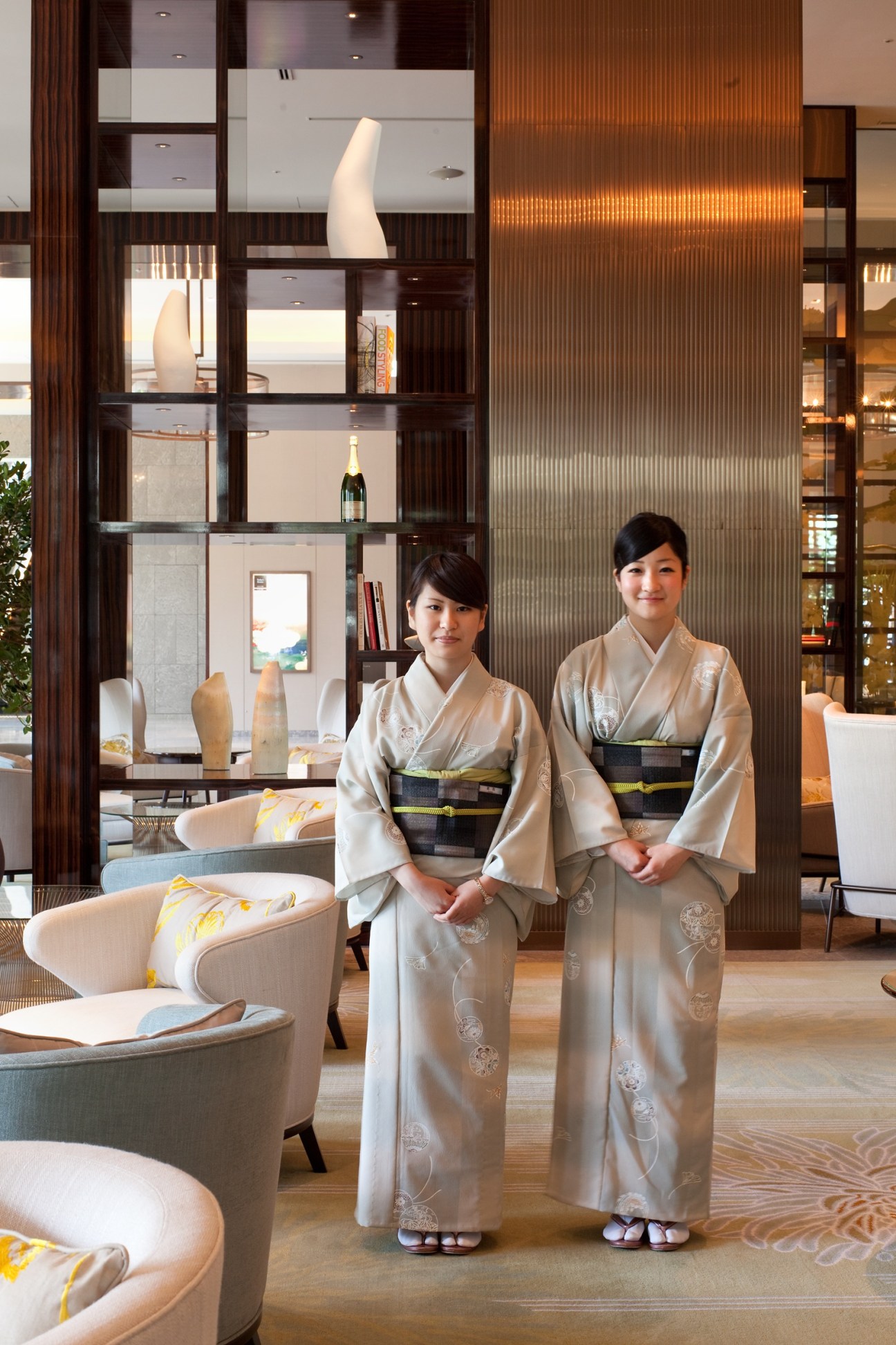 Two women in traditional Japanese kimonos stand in a modern hotel lobby with wooden lattice screening and contemporary furnishings.