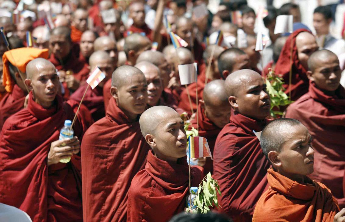 Buddhist monks in burgundy robes holding flags during a Myanmar political demonstration.