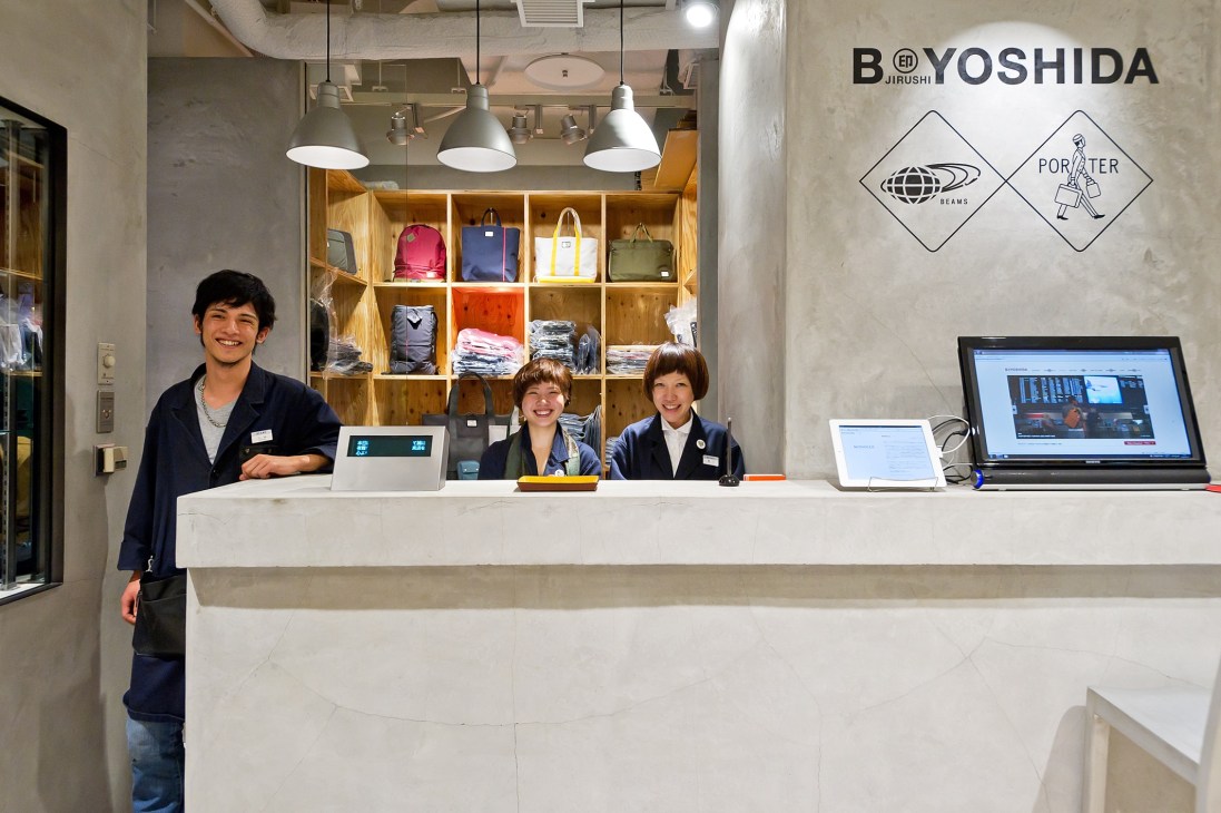 Bijirushi shop counter at Narita airport with three smiling staff members behind desk and wooden shelving displaying colorful backpacks and travel gear.