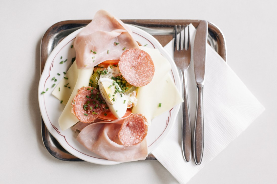 Open-faced sandwich with sliced salami, cheese, and pickles on a white plate with fork and knife.