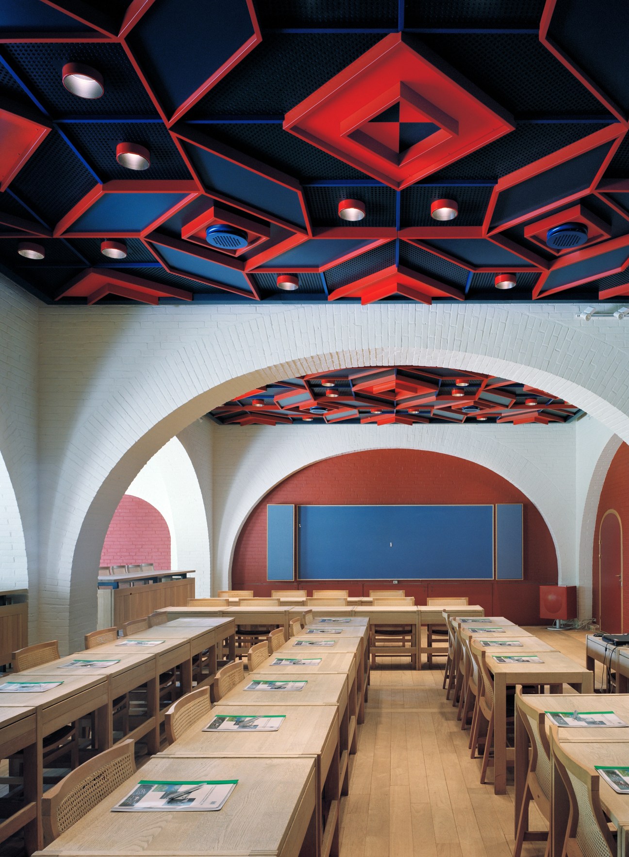 Lo-skolen's small conference hall with geometric red and blue ceiling reliefs, white arched walls, and wooden tables and chairs.