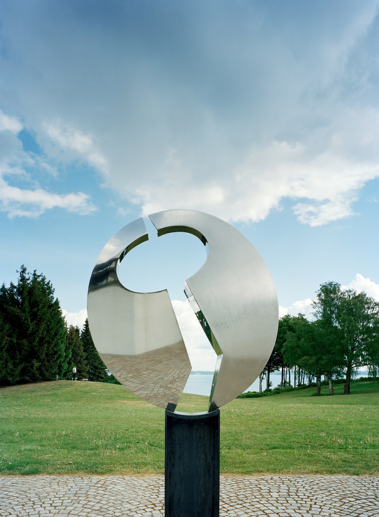 Abstract white metal sculpture by Børge Jørgensen on grassy grounds with trees and water view.