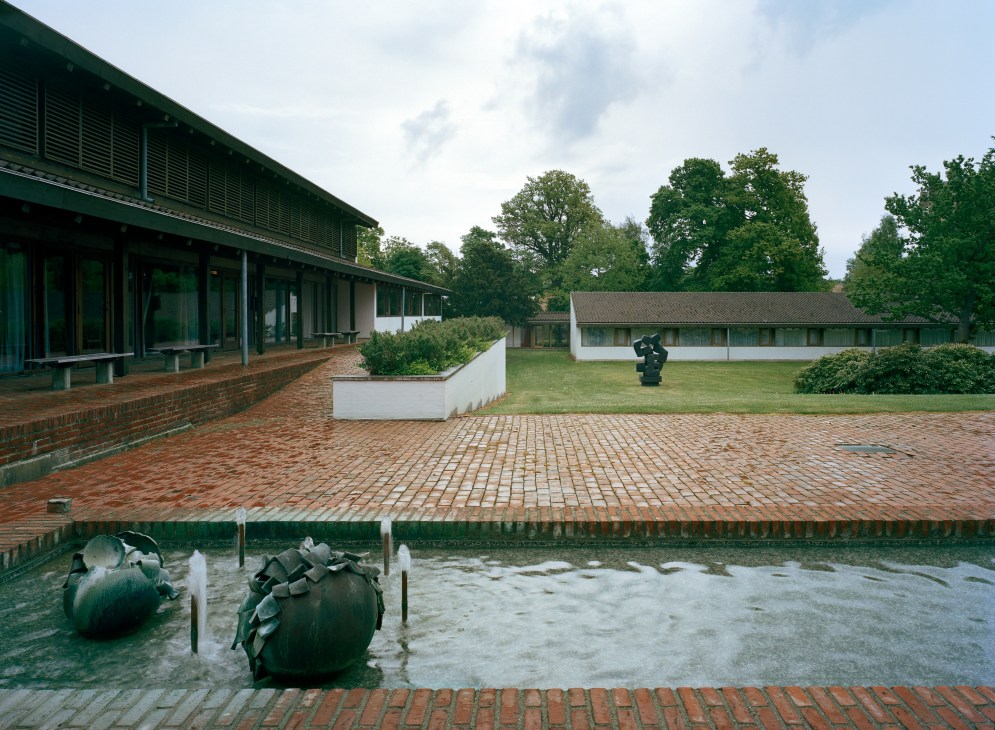 Lo-skolen campus courtyard with brick paving, modern building with green roof, white residential structures, and sculpture by water feature.