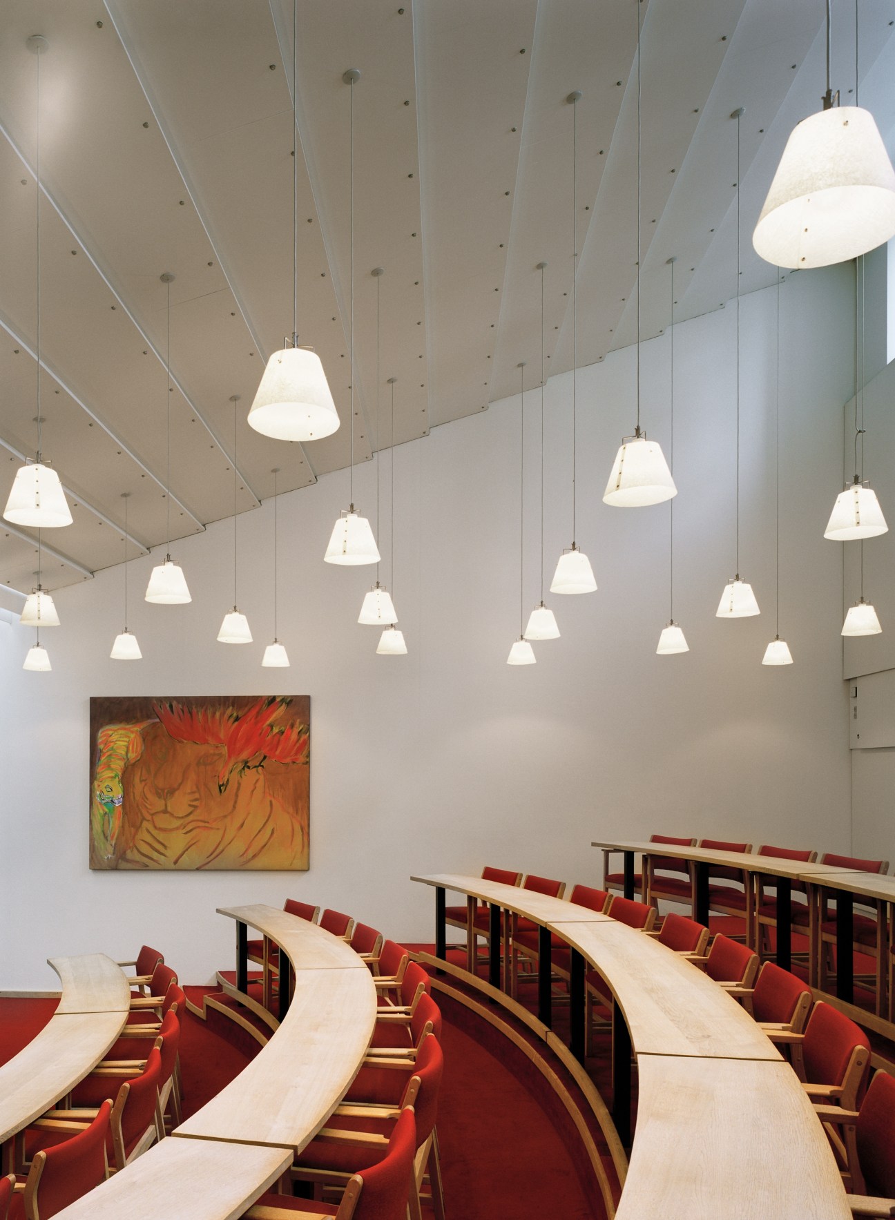 Lo-skolen's small auditorium with curved wooden seating, red upholstery, pendant lights, and a colorful abstract painting on the wall.
