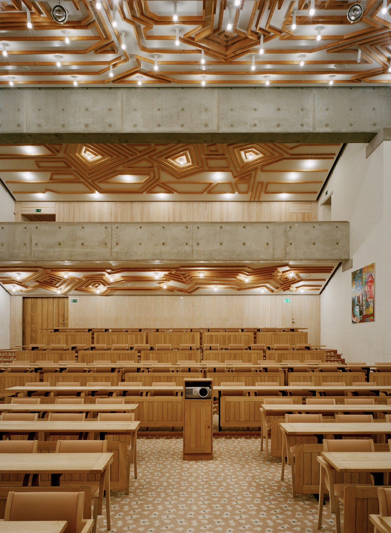 Lo-skolen conference hall interior with geometric wooden ceiling reliefs and rows of wooden chairs.