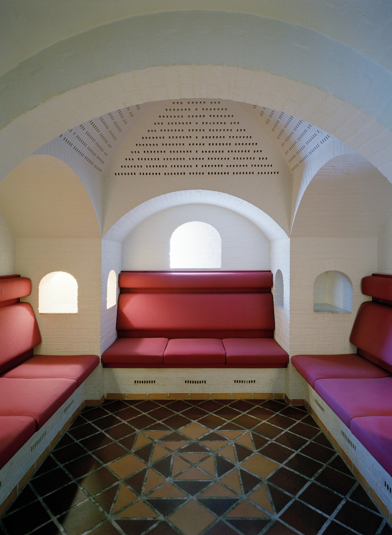 White-brick vaulted interior with red cushioned seating, geometric ceiling reliefs, and arched alcoves at Lo-skolen.
