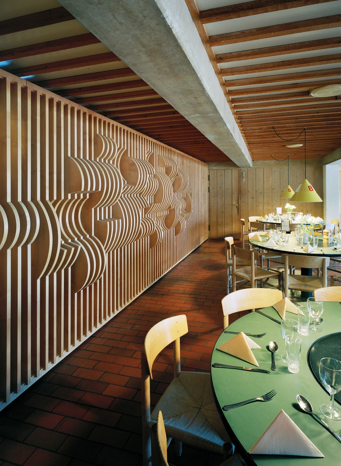 Lo-skolen restaurant with geometric patterned wall panel, wooden ceiling, and dining tables with modernist chairs.