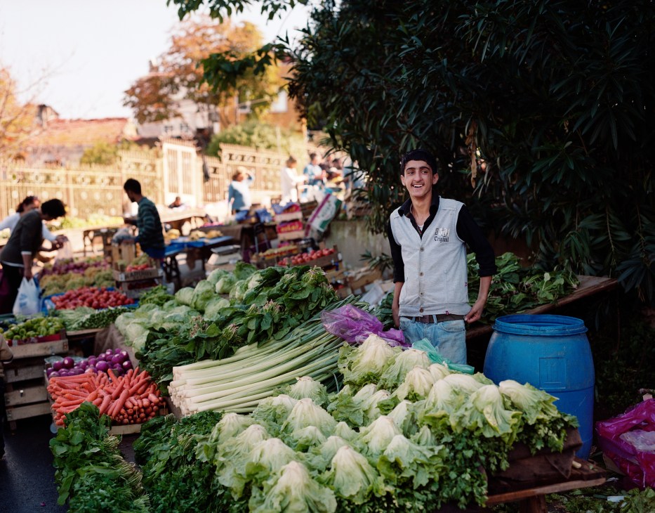 Fruitseller Mustafa Kemal stands smiling at a vegetable market stall displaying fresh produce.