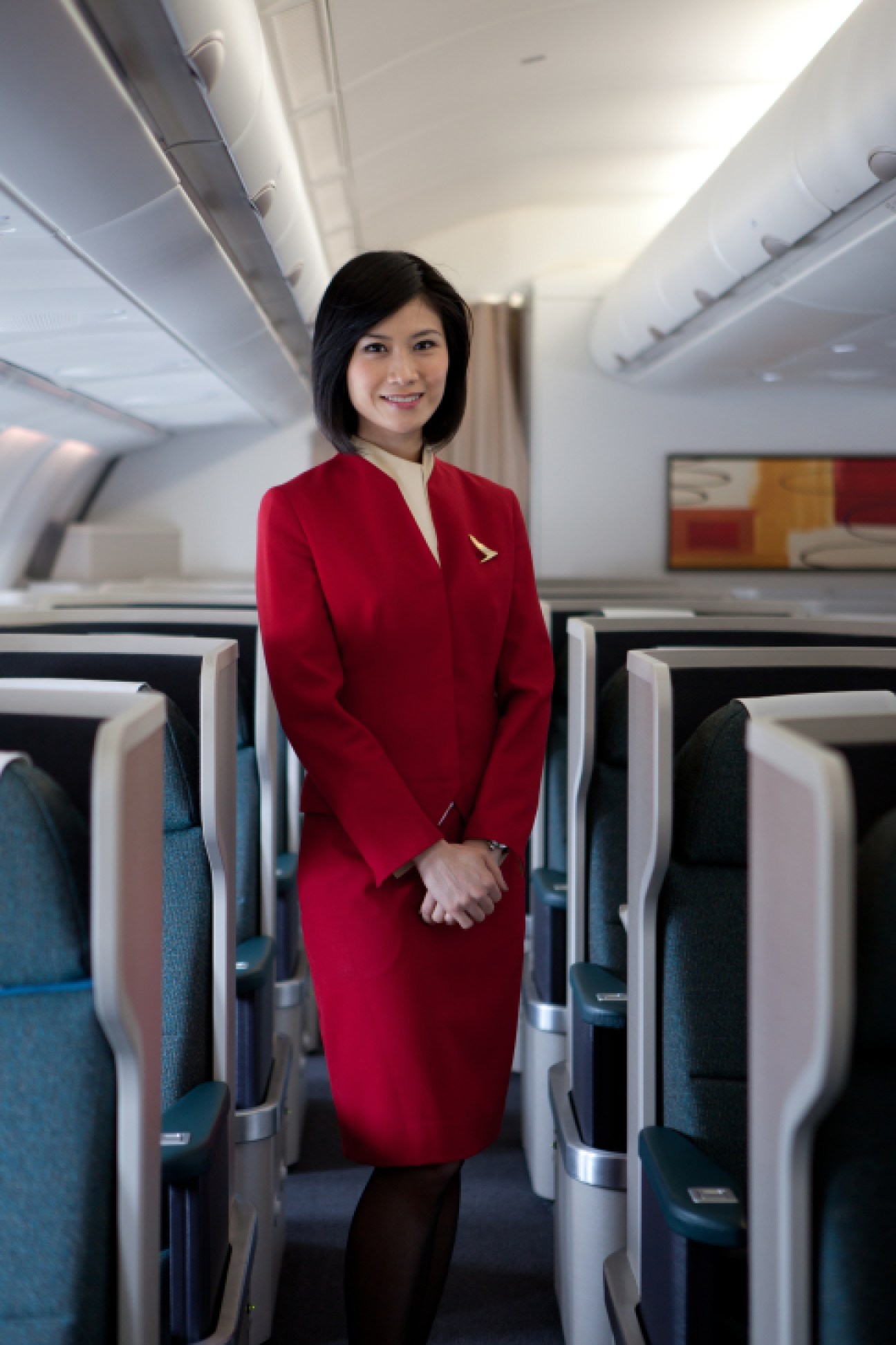 Cathay Pacific flight attendant in red uniform standing in aircraft cabin with business class seats.