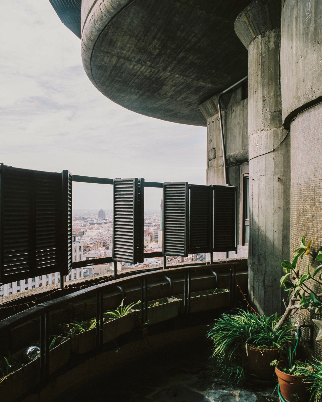 A circular balcony at Torres Blancas with black louvered screens, potted plants, and panoramic views of Madrid below.
