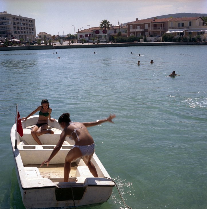 Young people swimming and playing in the sea near a white boat in Çesme, Turkey, with waterfront buildings visible in the background.