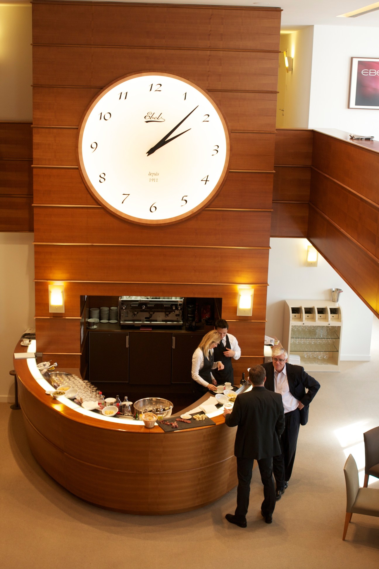 A large wall clock above a curved wooden bar counter where two people interact with a bartender.
