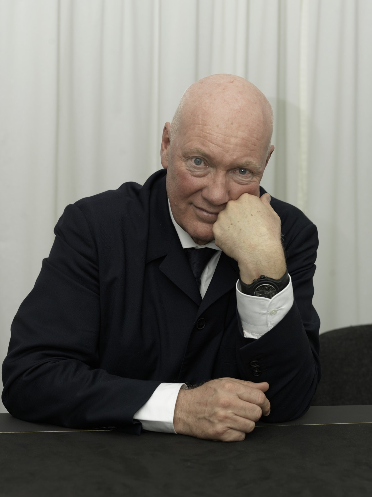 Jean-Claude Biver, CEO of Hublot, in a black suit and white shirt, leaning forward with his hand to his face at a table.