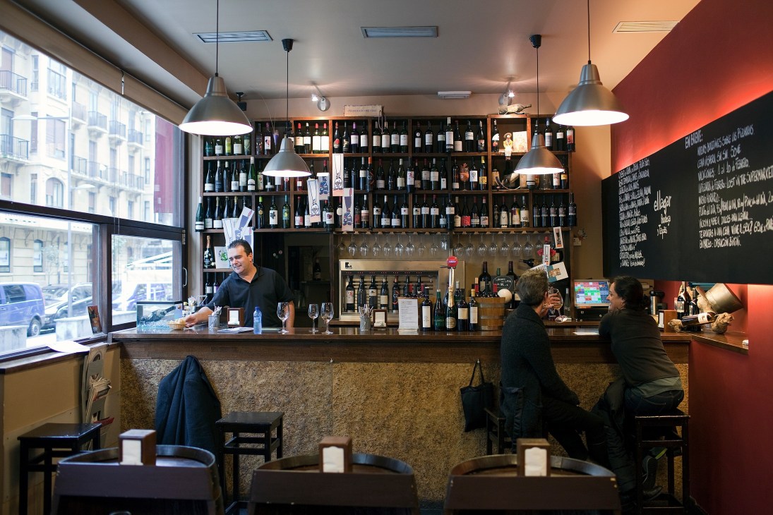 Interior of El Lagar pintxos bar with shelves of wine bottles and customers at the counter.