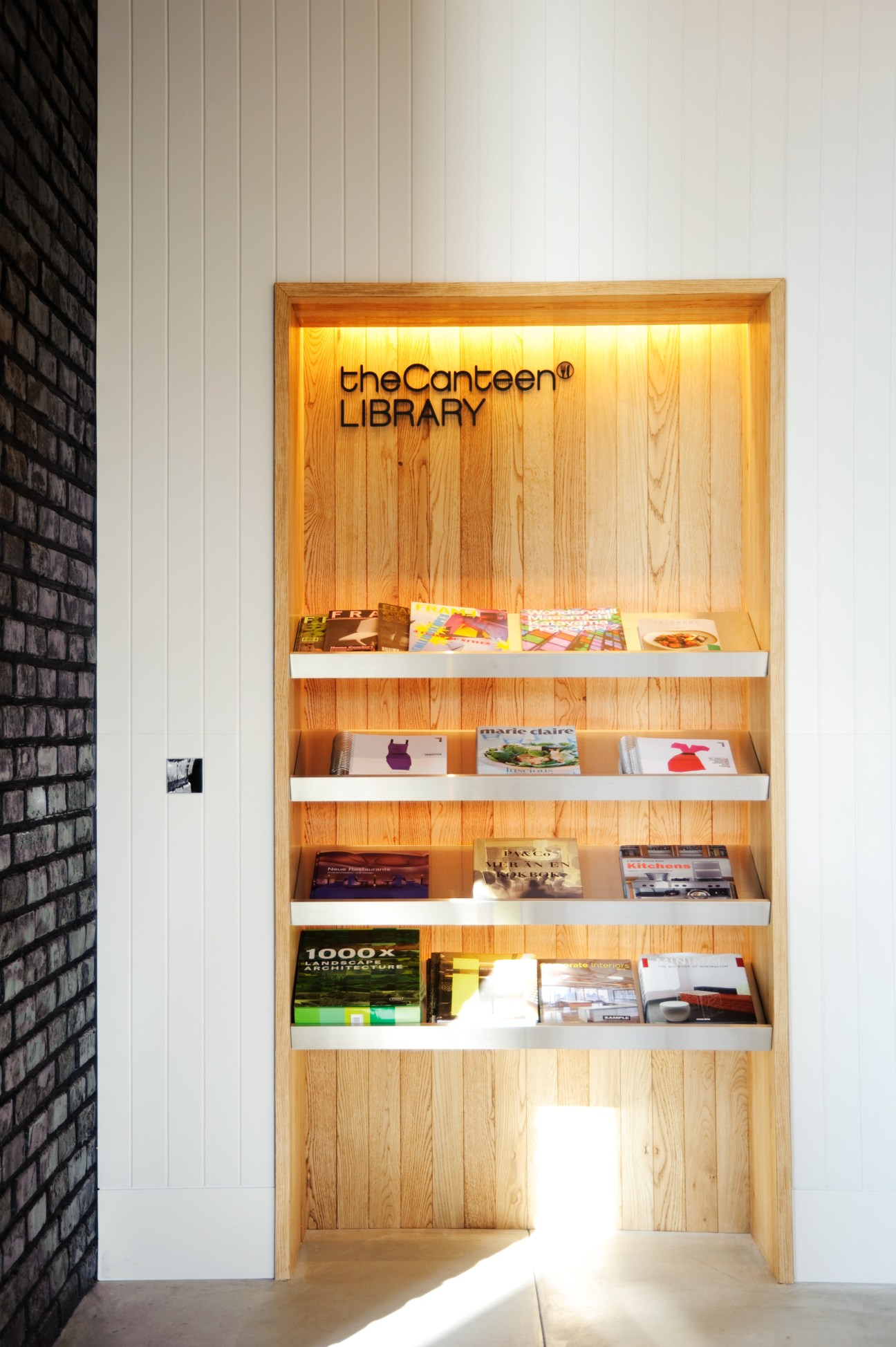 Wooden shelving unit displaying books and magazines with 'theCanteen LIBRARY' text and warm lighting.