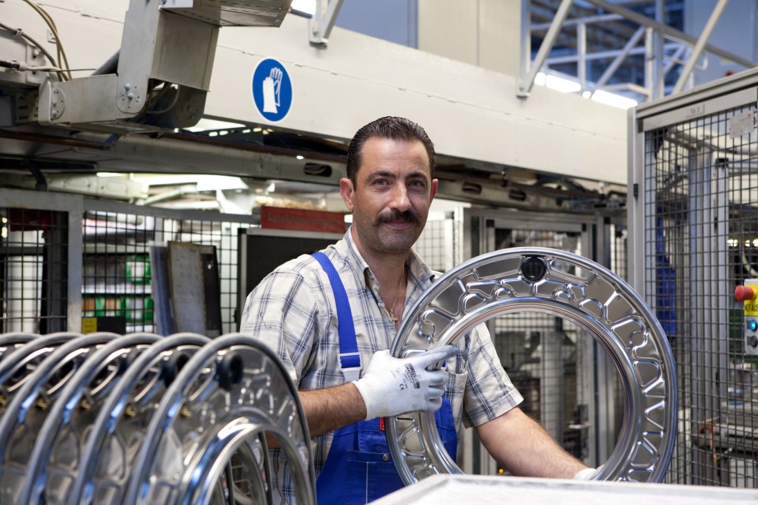 Factory worker in blue overalls and white gloves handling polished washing machine drums at Miele manufacturing facility.