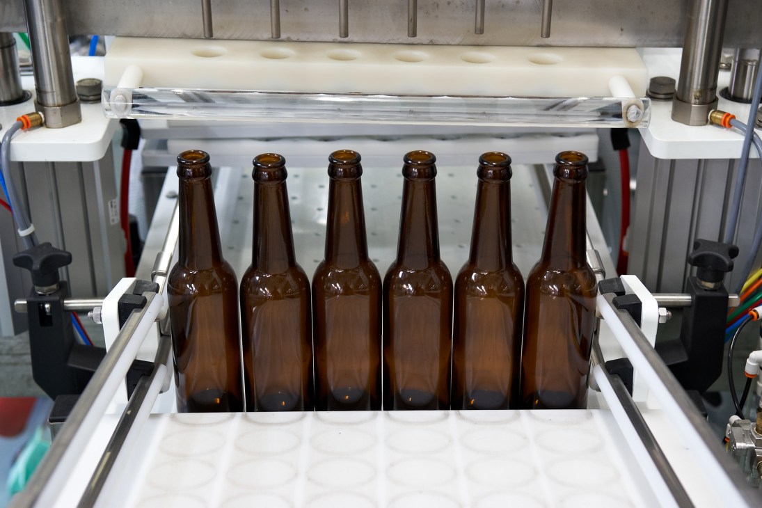 Six brown beer bottles arranged in a bottling machine with white and metal components.