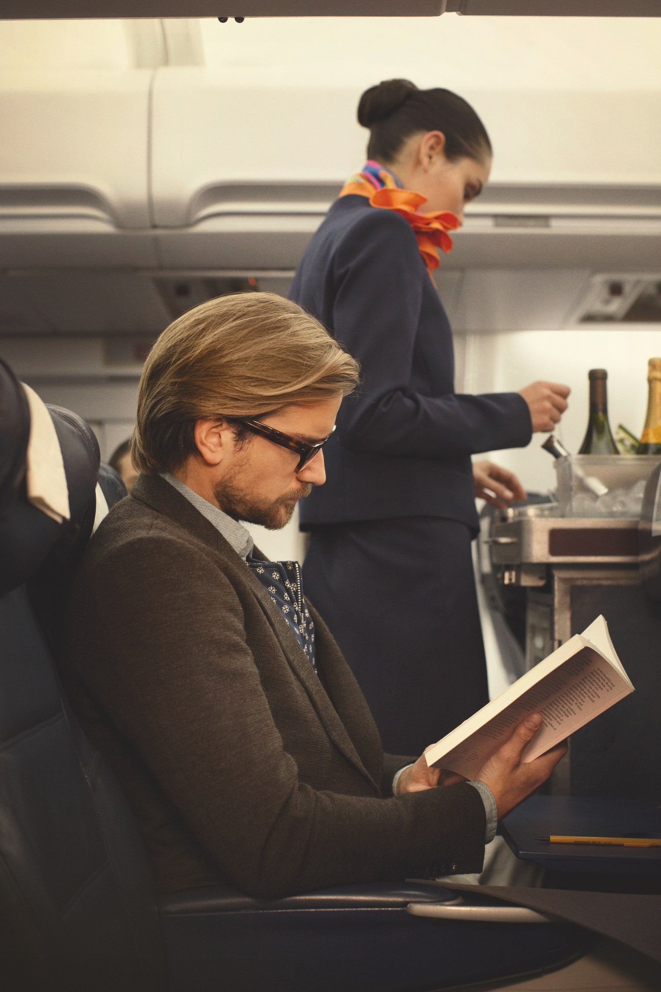 Man wearing Claire Goldsmith glasses and Strellson jacket reads on airplane with female passenger behind him.