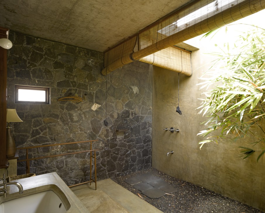 Open-air wet room with black basalt stone wall, concrete surfaces, and bamboo screening allowing natural light and vegetation visibility.