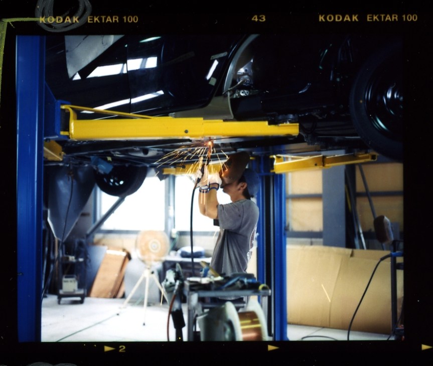 Worker welding car parts on a yellow overhead crane arm at Mitsuoka Motors factory.