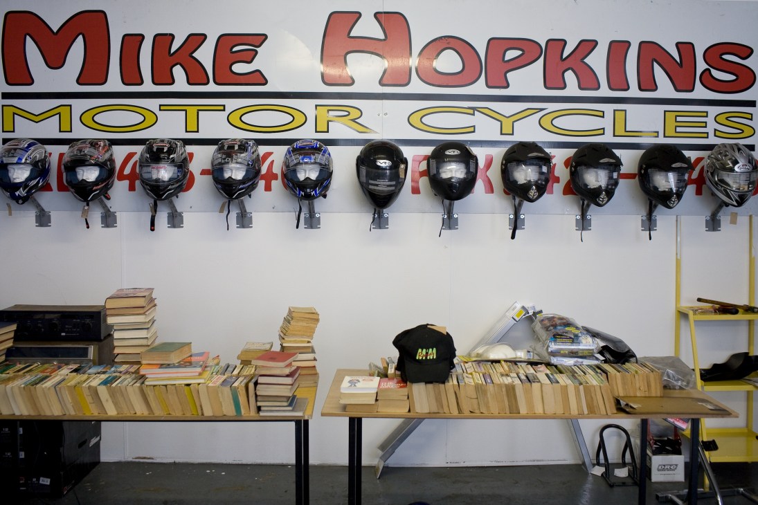 Mike Hopkins Motorcycles storefront with red and yellow signage, motorcycle helmets on wall, and books displayed on tables below.