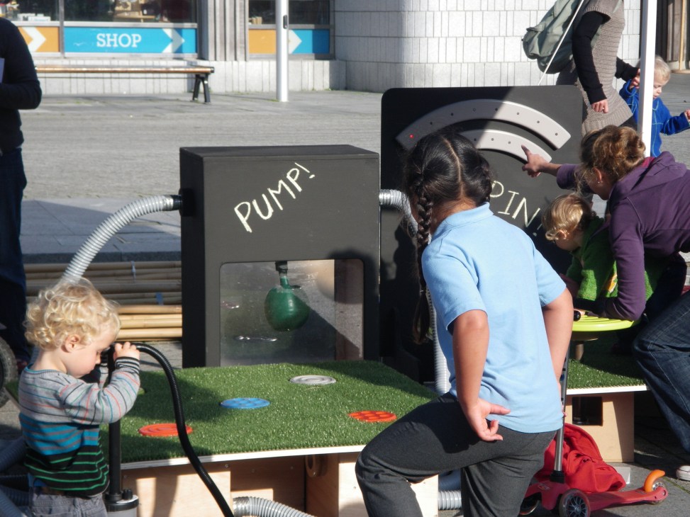 Children interact with an energy-harnessing playground installation featuring pump and spinning mechanisms.