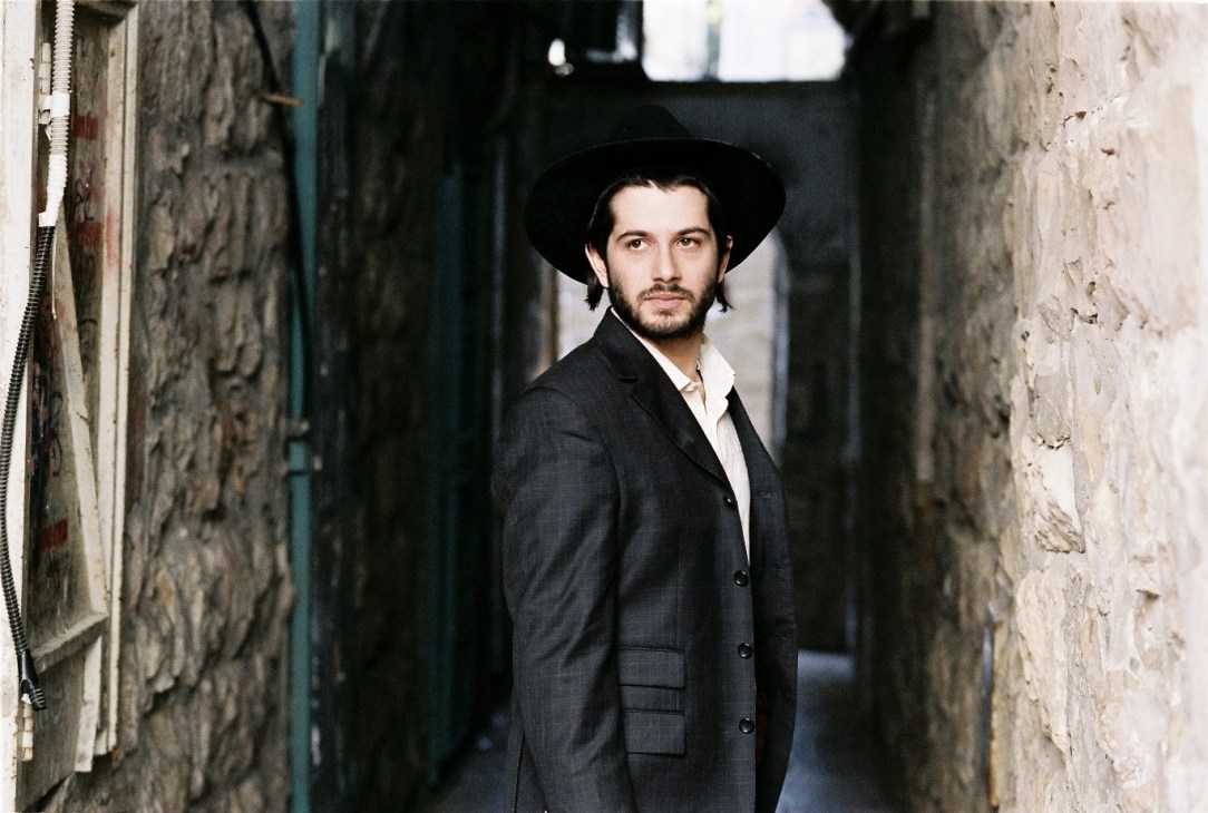 Man in dark suit and black hat standing in narrow alley between weathered stone walls.