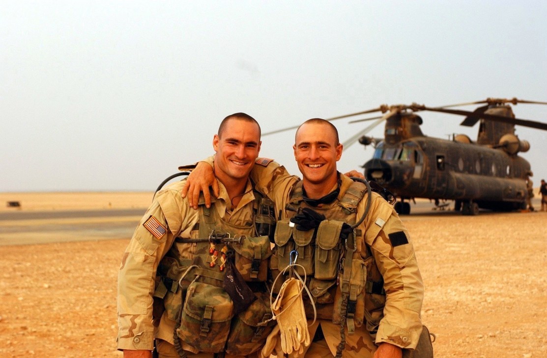 Two US soldiers in desert camouflage uniforms smile at the camera with a military helicopter behind them in an arid landscape.