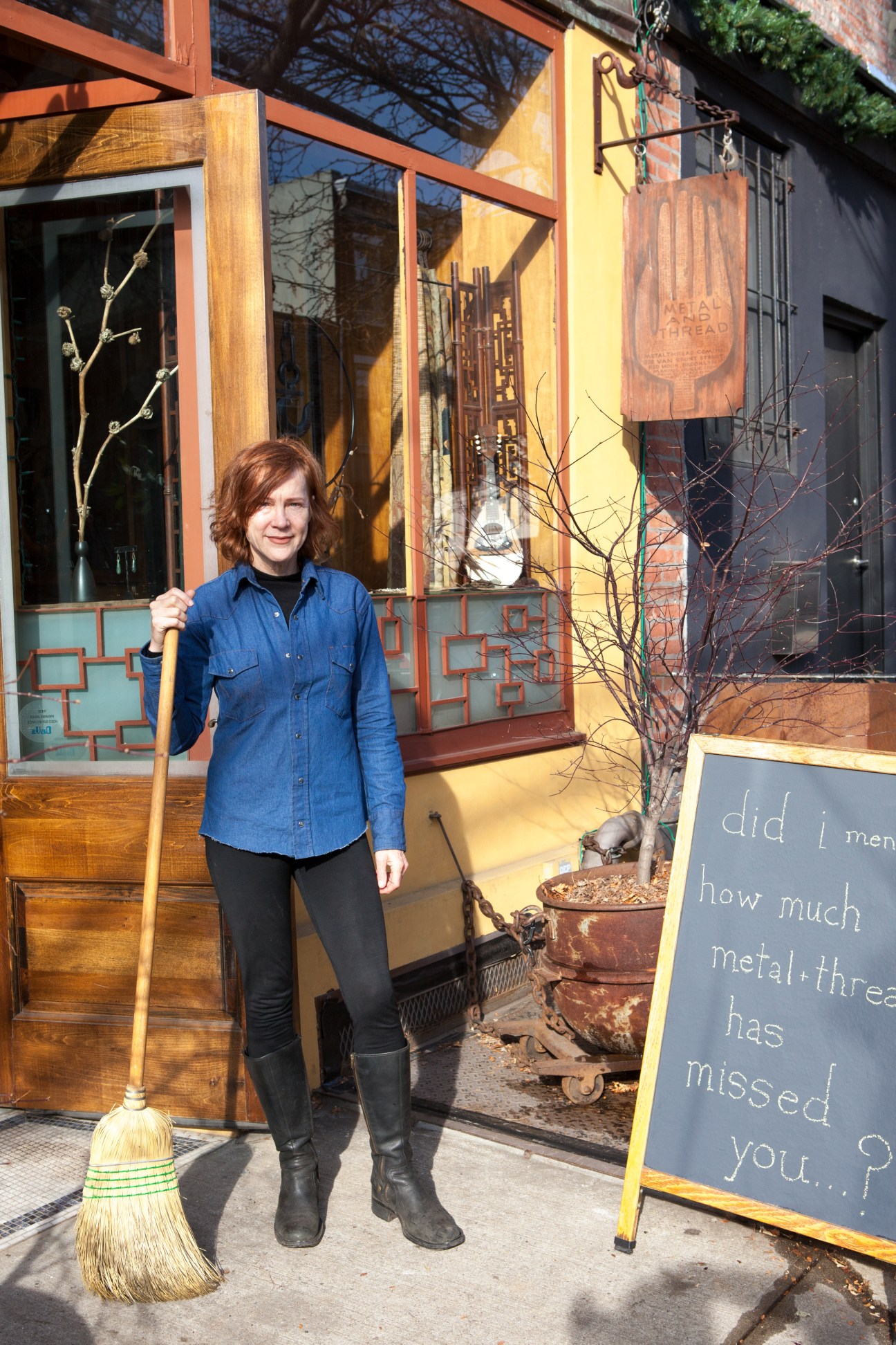 Woman in blue shirt holding a broom stands outside Metal and Thread storefront in Red Hook, Brooklyn