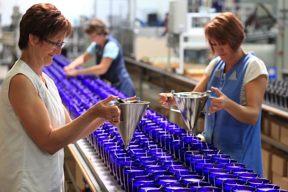 Factory workers pouring melted wax into blue glass jars on a production line.